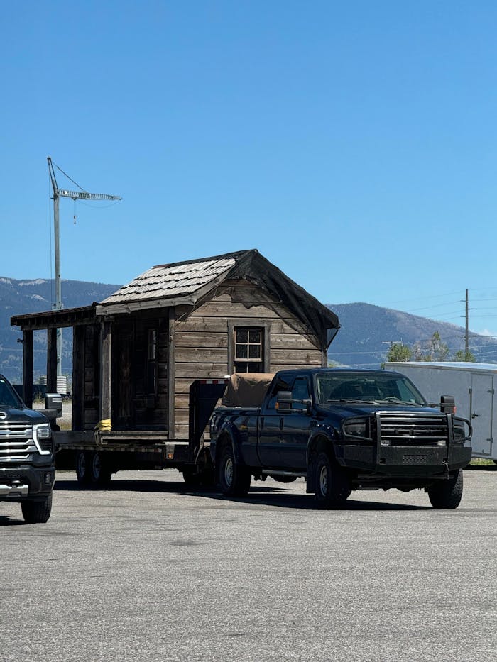 A rustic wooden cabin being towed by a pickup truck on a clear, sunny day.